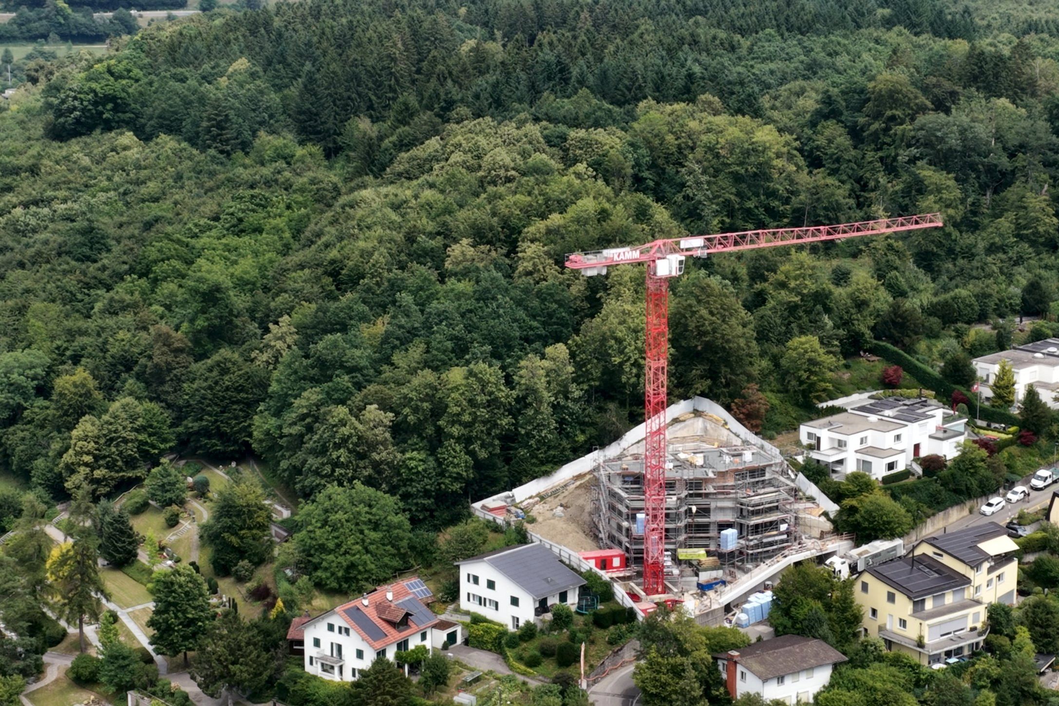 Drohnenbild: eingerüsteter Rohbau mit rotem Turmdrehkran am Waldrand, umgeben von Wohnhäusern