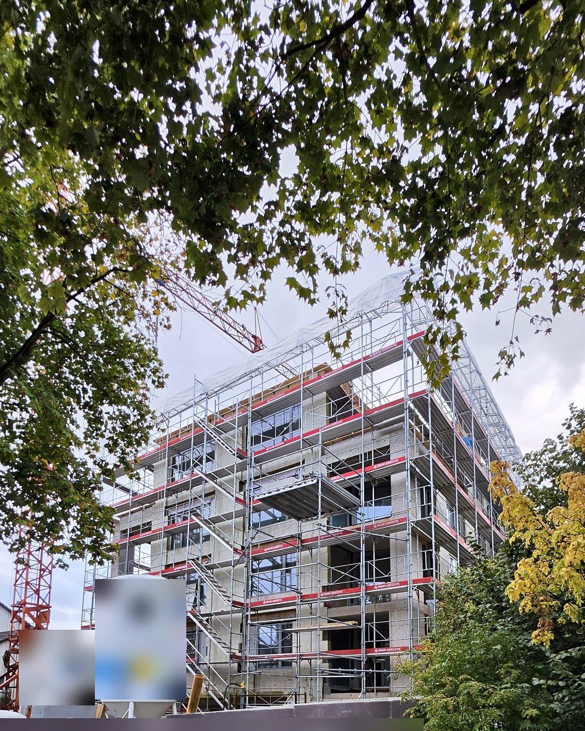 Eingerüsteter Bau im Oktober hinter Bäumen, Tafeln und Silo unkenntlich gemacht